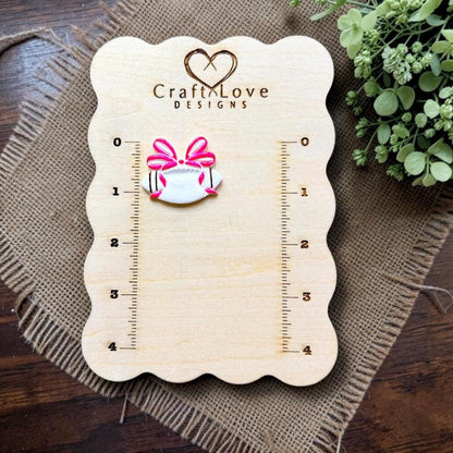 Finished football clay piece with a bright pink oversized bow displayed on a Craft Love Designs measurement board for scale.