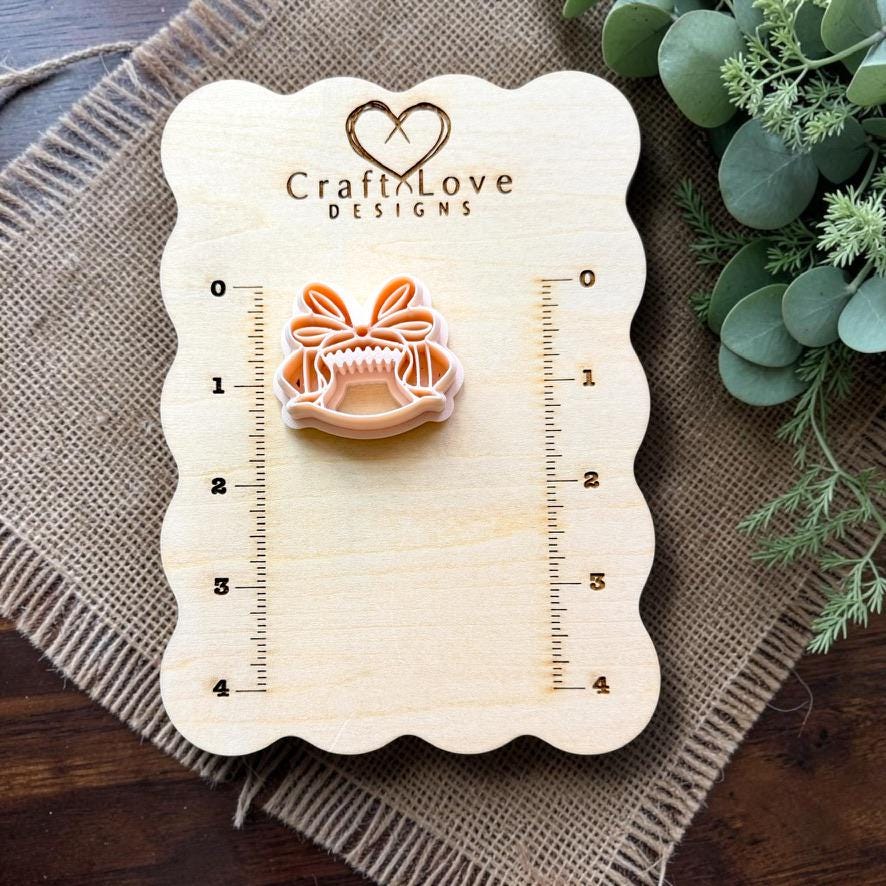 Coquette bow football polymer clay cutter displayed on a Craft Love Designs measurement board for size reference.