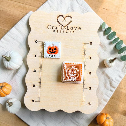 Framed jack-o-lantern pumpkin clay piece and matching polymer clay cutter displayed on a measurement board with autumn pumpkins nearby.