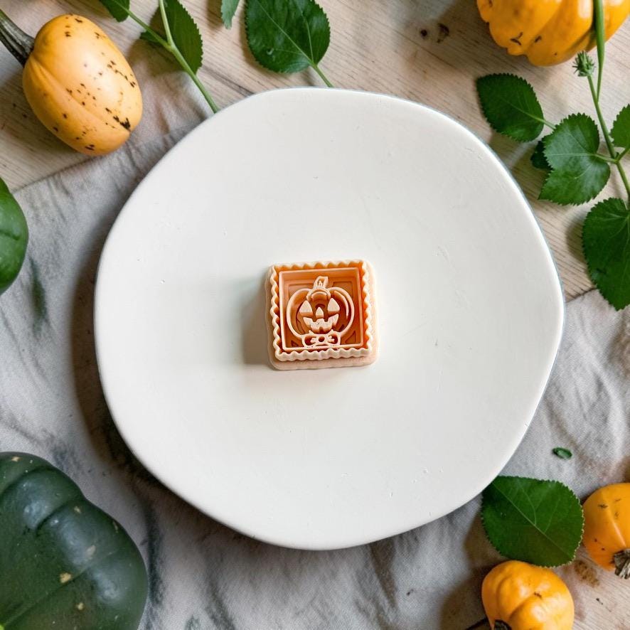 Framed jack-o-lantern pumpkin polymer clay cutter displayed on a white plate surrounded by autumn pumpkins and greenery.