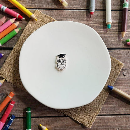 Finished white owl wearing a black graduation cap displayed on a white plate surrounded by colorful pencils, crayons, and school supplies.