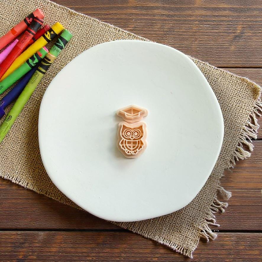 Two-piece graduation owl polymer clay cutter with separate owl and graduation cap designs displayed on a white plate with colorful crayons nearby.