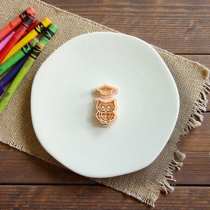 Two-piece graduation owl polymer clay cutter with separate owl and graduation cap designs displayed on a white plate with colorful crayons nearby.