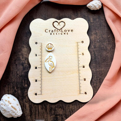 Finished pelican and seashell topper polymer clay earrings displayed on a CraftyLove Designs sizing board with coastal décor.