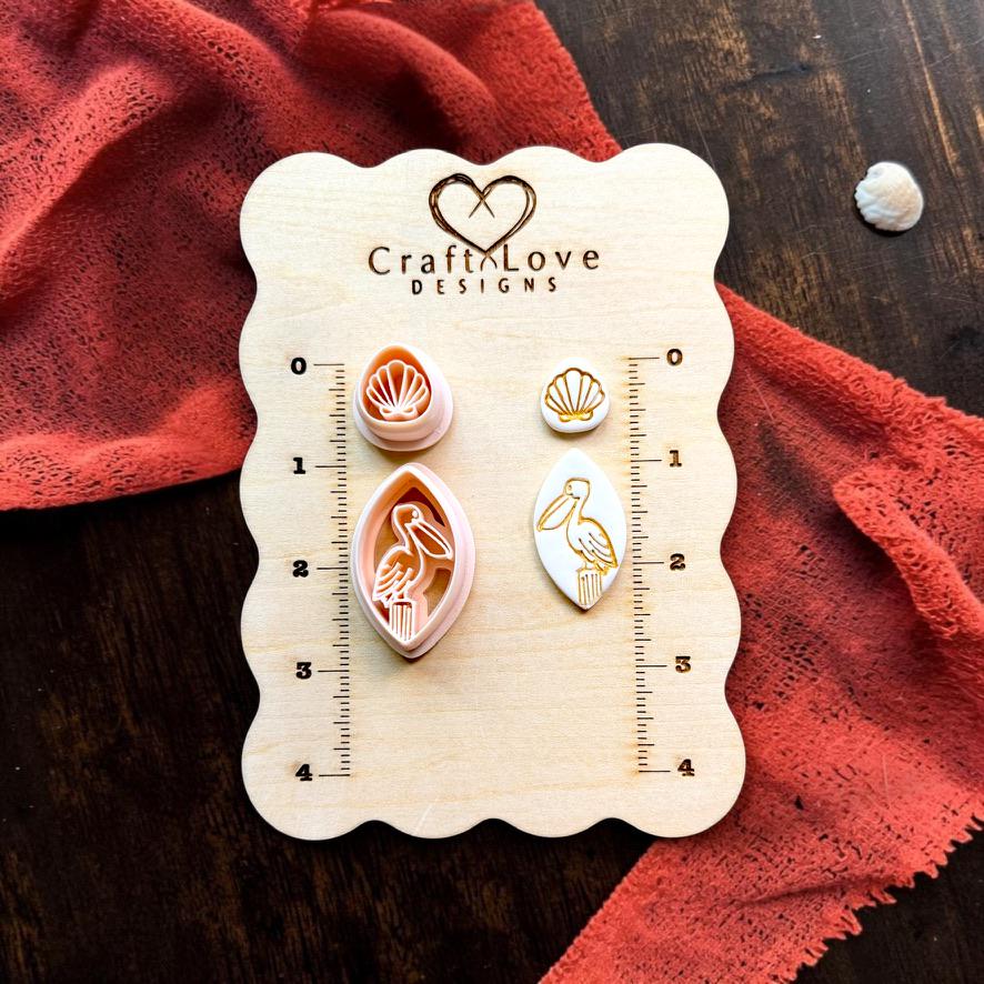 Pelican polymer clay cutter and matching seashell topper cutter displayed beside finished clay pieces on a CraftyLove Designs sizing board.