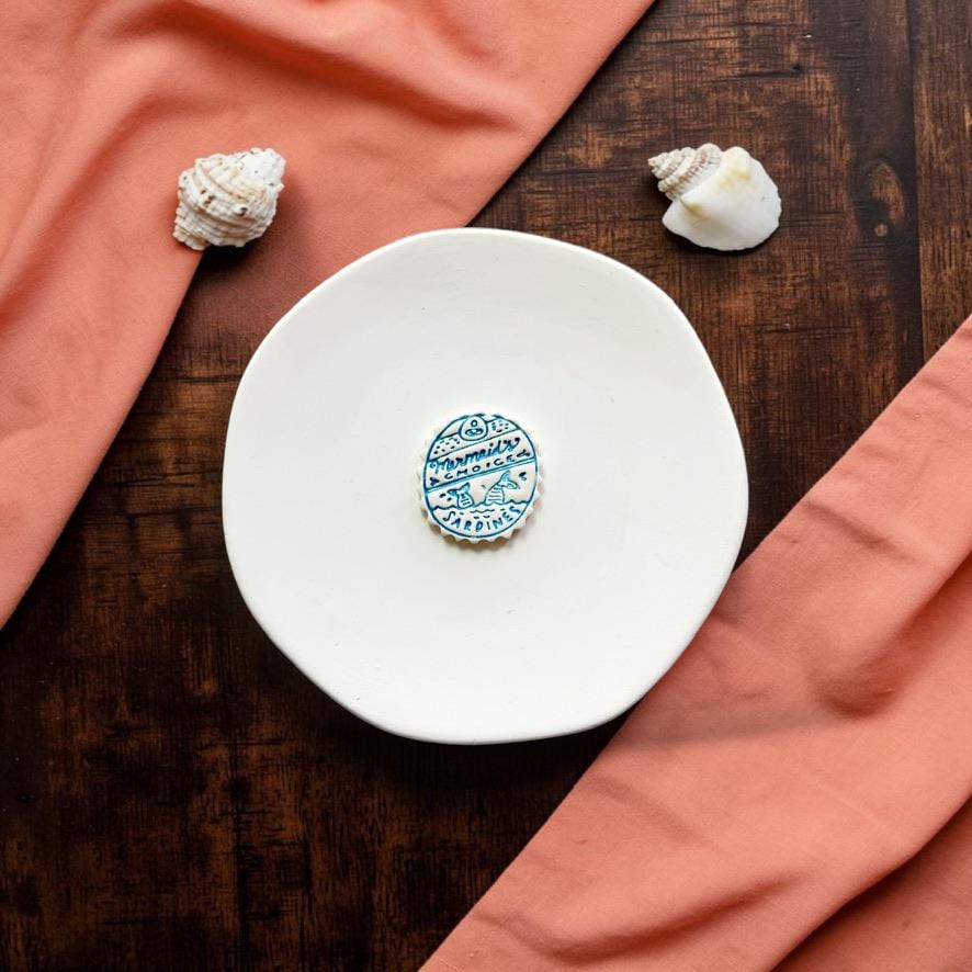 Finished Mermaid Sardines clay design displayed on a white ceramic plate with seashells and coral fabric.