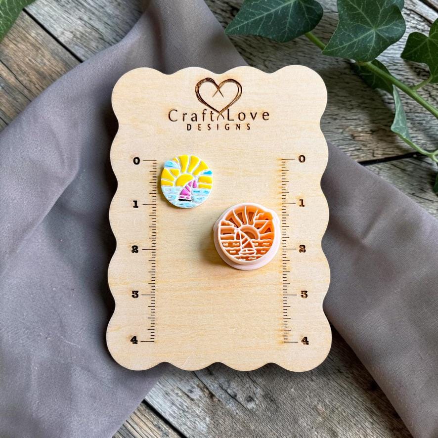 Finished colorful sailboat sunset polymer clay earring displayed beside the matching cutter on a CraftyLove Designs sizing board.