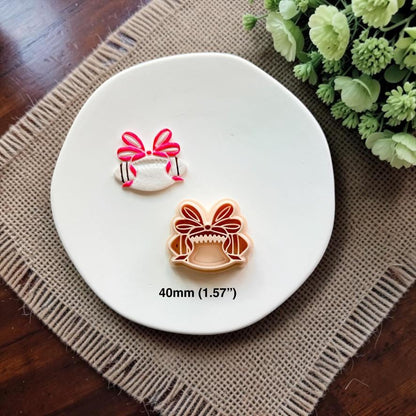 Finished white football clay piece with a bright pink coquette bow beside the matching 40mm polymer clay cutter.