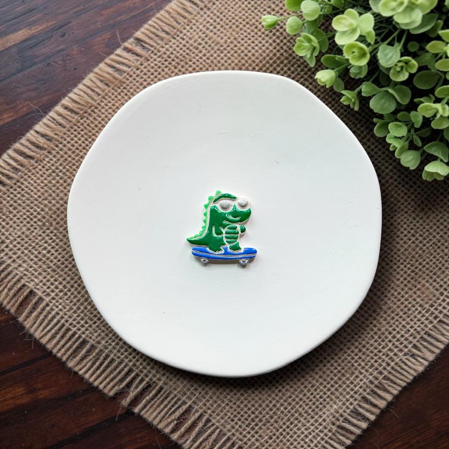 Finished green gator wearing silver sunglasses and riding a blue skateboard, displayed on a white plate.