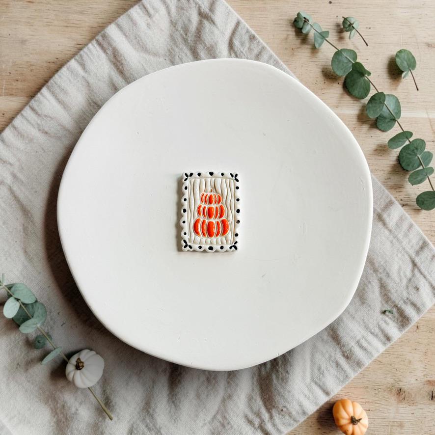 Finished white polymer clay piece featuring three stacked orange pumpkins, textured vertical details, and a decorative black border.