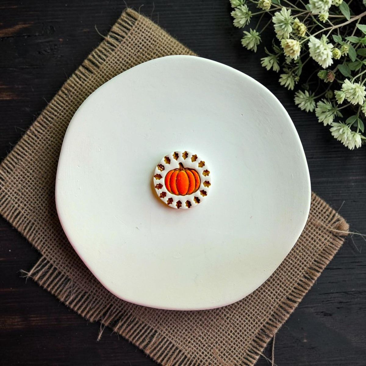 Finished white clay pumpkin earring with orange pumpkin and autumn leaf border displayed on a white dish.