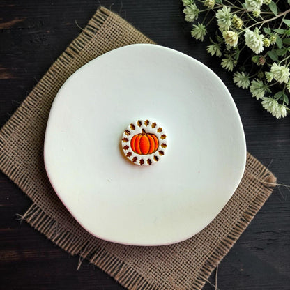 Finished white clay pumpkin earring with orange pumpkin and autumn leaf border displayed on a white dish.