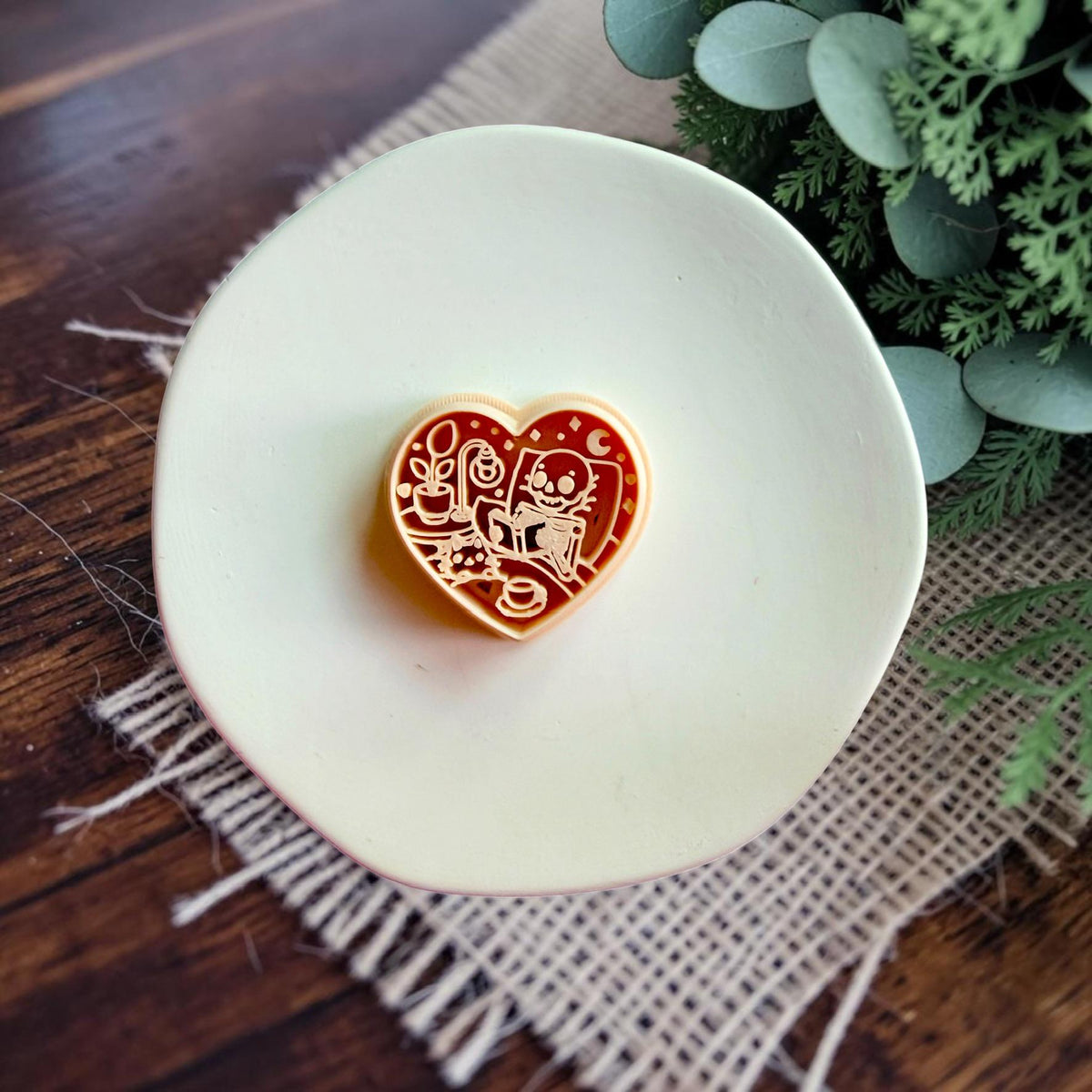 Heart-shaped polymer clay cutter featuring a cozy skeleton reading a book beside a lamp and houseplant.