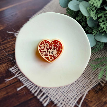 Heart-shaped polymer clay cutter featuring a cozy skeleton reading a book beside a lamp and houseplant.