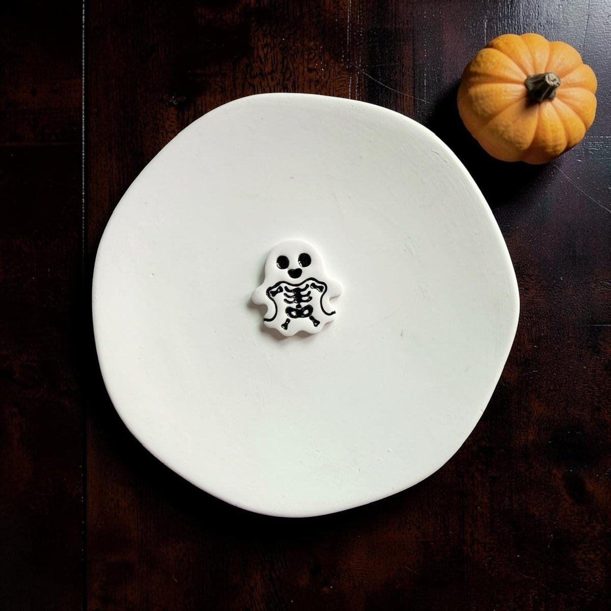 Finished white clay skeleton ghost with black eyes and detailed black ribcage centered on a white round surface beside a small orange pumpkin.