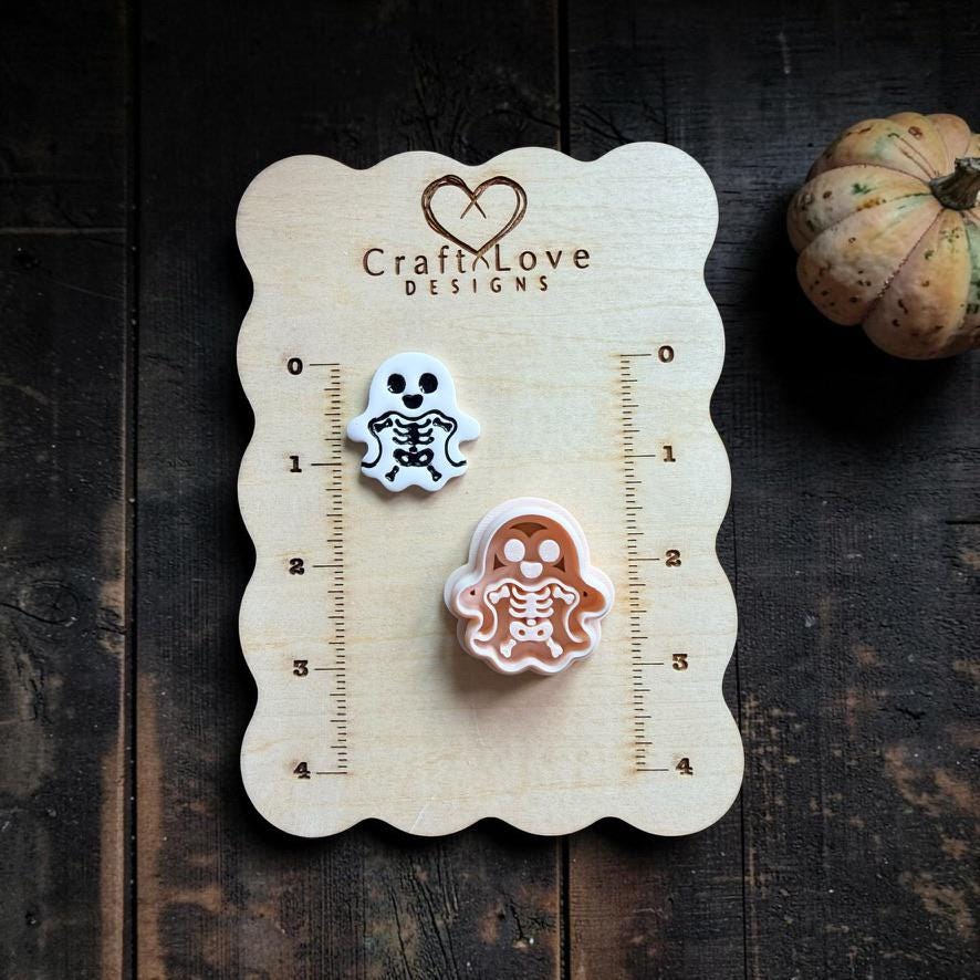Finished white skeleton ghost clay piece beside the matching peach-colored cutter on a wooden Craft Love Designs ruler board showing their scale.