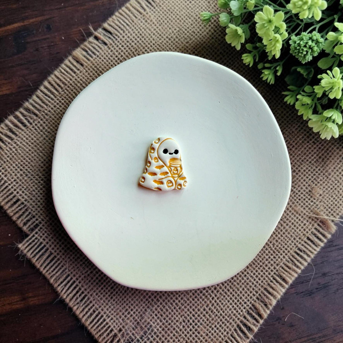Finished polymer clay ghost wrapped in a blanket holding a coffee cup displayed on a white dish.