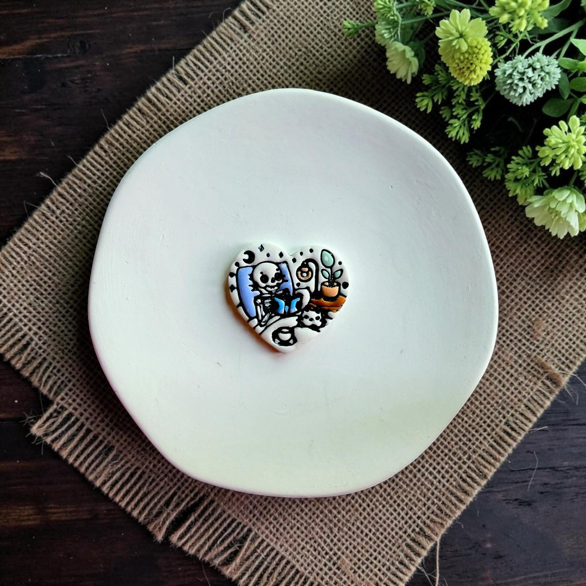 Finished heart-shaped polymer clay design featuring a cozy skeleton reading beneath a moon beside a lamp and plant.