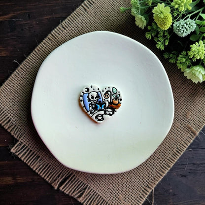 Finished heart-shaped polymer clay design featuring a cozy skeleton reading beneath a moon beside a lamp and plant.