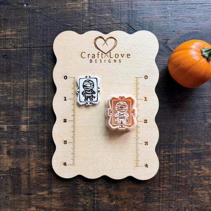 Finished white clay mummy piece and peach mummy design displayed on a Craft Love Designs wooden measuring board, showing a wrapped mummy character inside a decorative rectangular frame.