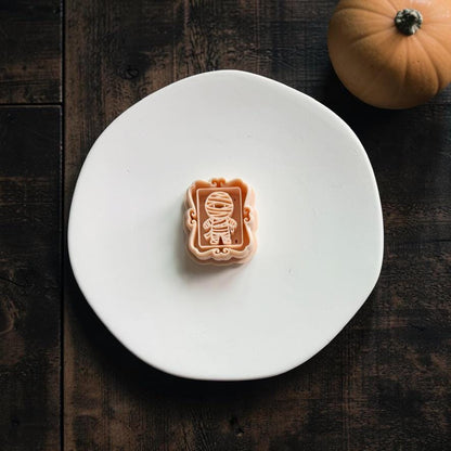 Peach framed mummy design centered on a large white clay slab over a dark wood background, with a small orange pumpkin in the upper right corner.