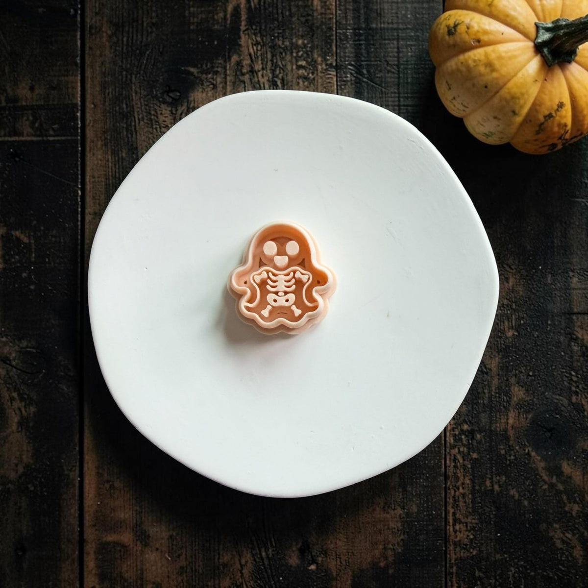 Skeleton ghost polymer clay cutter centered on a white round surface with a small pumpkin and dark wood Halloween background.