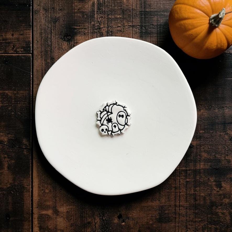 Black-and-white example clay piece featuring ghost, skull, spider and web details on a white plate with a pumpkin in the background.