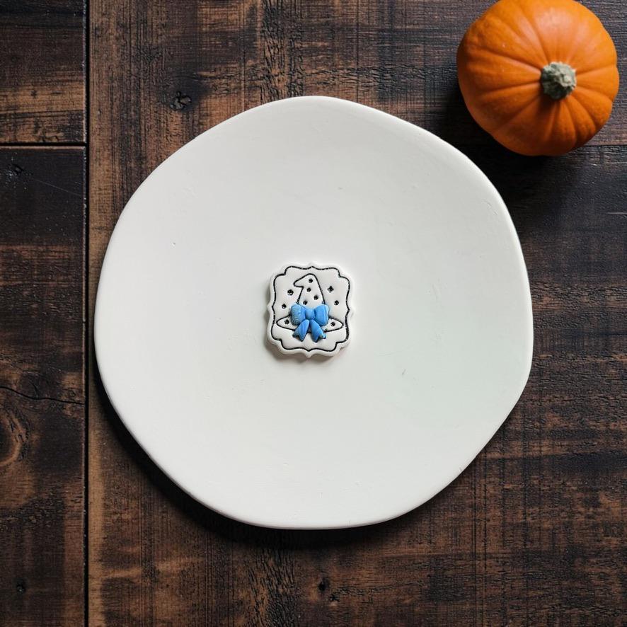 Finished white clay piece centered on a white clay slab, featuring a pointed witch hat with stars and a bright blue bow, with an orange pumpkin in the upper right.
