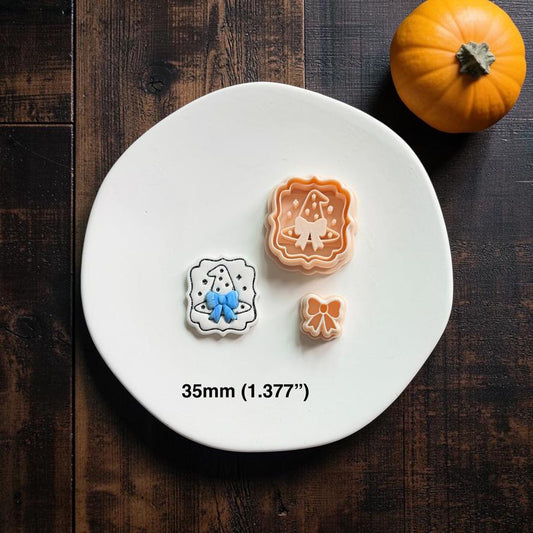 Finished white witch hat piece with blue bow shown beside the peach main design and separate bow on a white clay slab, with an orange pumpkin in the upper right.