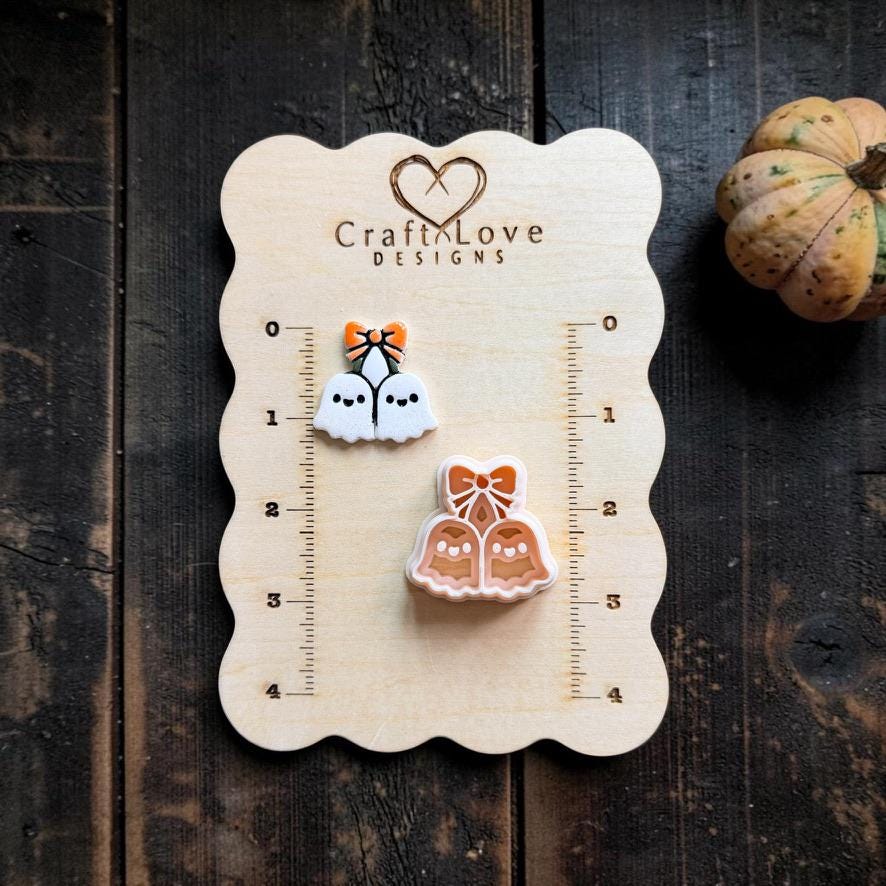 Finished white ghost cherries with black faces and an orange bow displayed beside the matching peach design on a Craft Love Designs wooden measuring board.