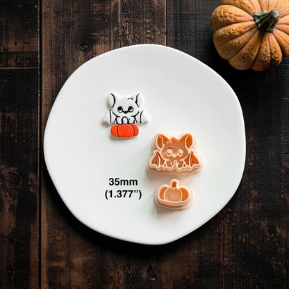 Finished white bat perched on an orange pumpkin beside the matching peach bat and separate pumpkin designs on a white clay plate.