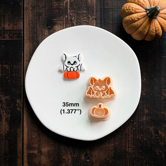 Finished white bat perched on an orange pumpkin beside the matching peach bat and separate pumpkin designs on a white clay plate.