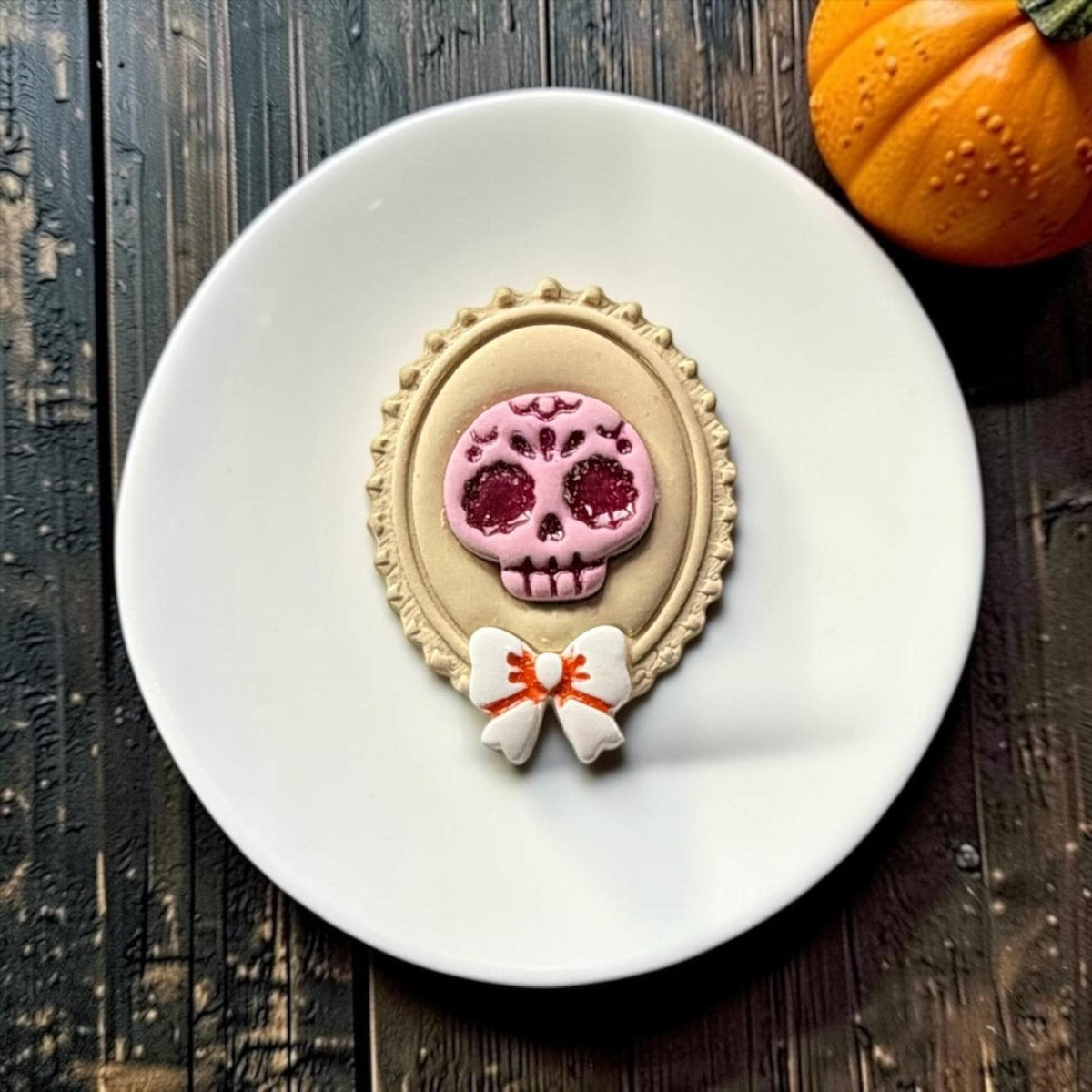 Close-up of the finished sugar skull cameo on a white round surface, featuring a pink decorated skull with dark red glitter details, cream ornate oval frame, and white bow with orange accent.
