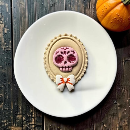 Close-up of the finished sugar skull cameo on a white round surface, featuring a pink decorated skull with dark red glitter details, cream ornate oval frame, and white bow with orange accent.
