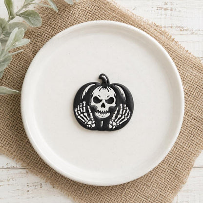 Finished black pumpkin clay piece displayed on a white plate, featuring a white skull in the center and detailed white skeleton hands on both sides.