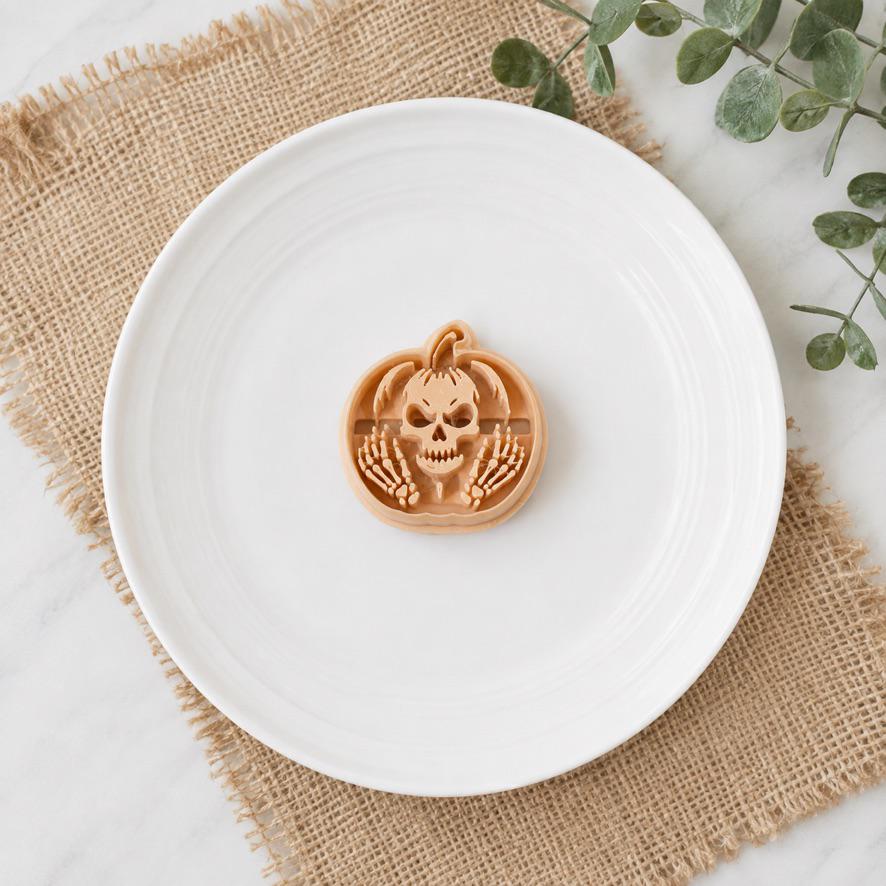 Peach 40mm skeleton pumpkin polymer clay cutter centered on a white plate, showing a skull surrounded by skeletal hands inside the pumpkin shape.
