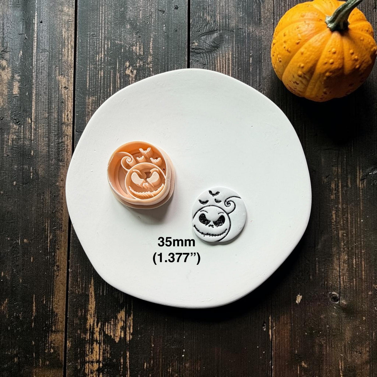 Peach 35mm pumpkin face clay cutter beside a finished white and black clay piece on a white plate, showing a grinning face, curled stem, and three flying bats.
