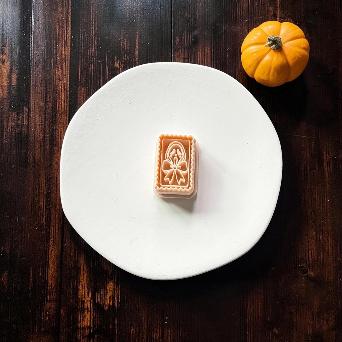 Peach rectangular clay cutter with a scalloped border, spooky elongated ghost face, hood outline, and large bow, centered on a white plate with a pumpkin nearby.