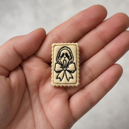 Close-up of a finished cream-colored ghost face and bow clay piece resting in a hand, showing the black facial details, oversized bow, rectangular frame, and scalloped edge.