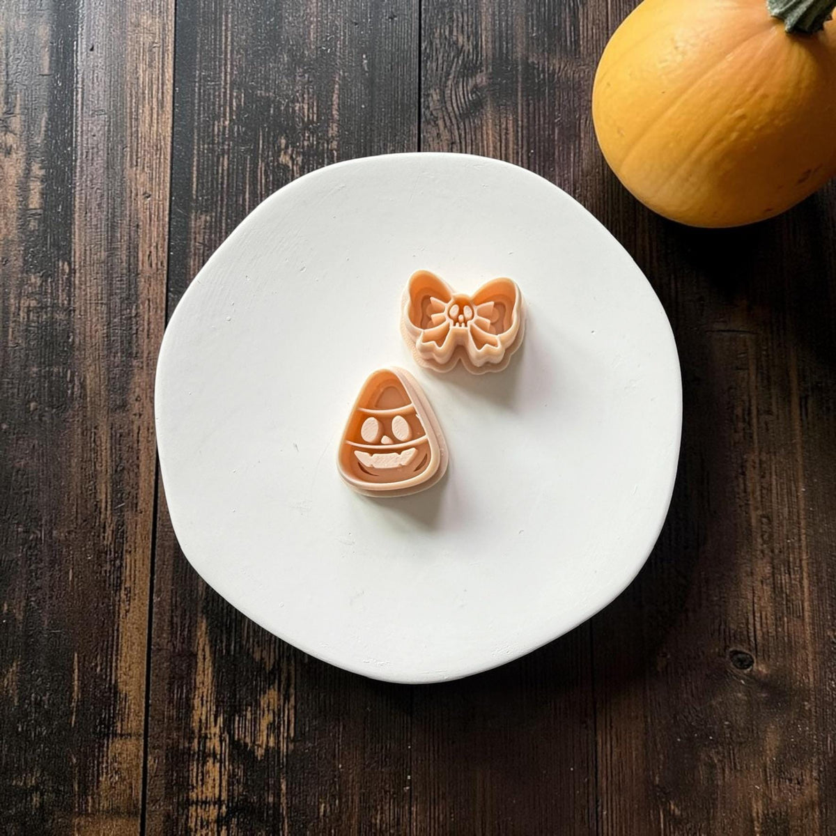 Peach two-piece clay design on a white plate featuring a smiling candy corn skull shape and a separate oversized bow with a tiny skull in the center, with a pumpkin in the background.