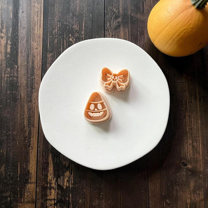 Peach two-piece clay design on a white plate featuring a smiling candy corn skull shape and a separate oversized bow with a tiny skull in the center, with a pumpkin in the background.