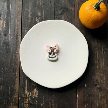 Finished Halloween clay piece centered on a white plate, featuring a white smiling candy corn skull face with dark glitter details and a pale pink bow topped with a tiny skull.