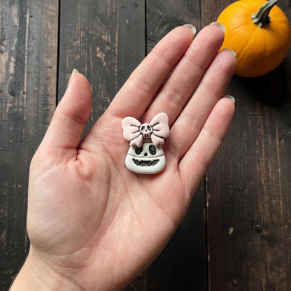 Finished Halloween clay piece centered on a white plate, featuring a white smiling candy corn skull face with dark glitter details and a pale pink bow topped with a tiny skull on hand.