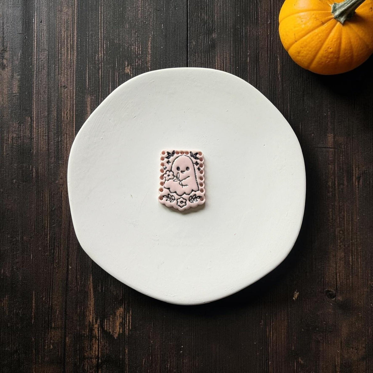 Finished pale pink rectangular clay piece centered on a white plate, featuring a cute ghost holding flowers with black detailed bats, stars, blossoms, leaves, and decorative edging.