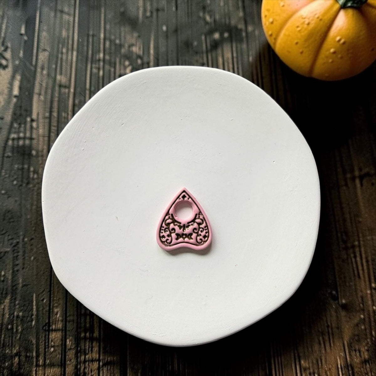 Finished pink planchette-shaped clay piece with contrasting dark details centered on a white plate, showing a circular opening, crescent moon, and intricate gothic ornamentation.
