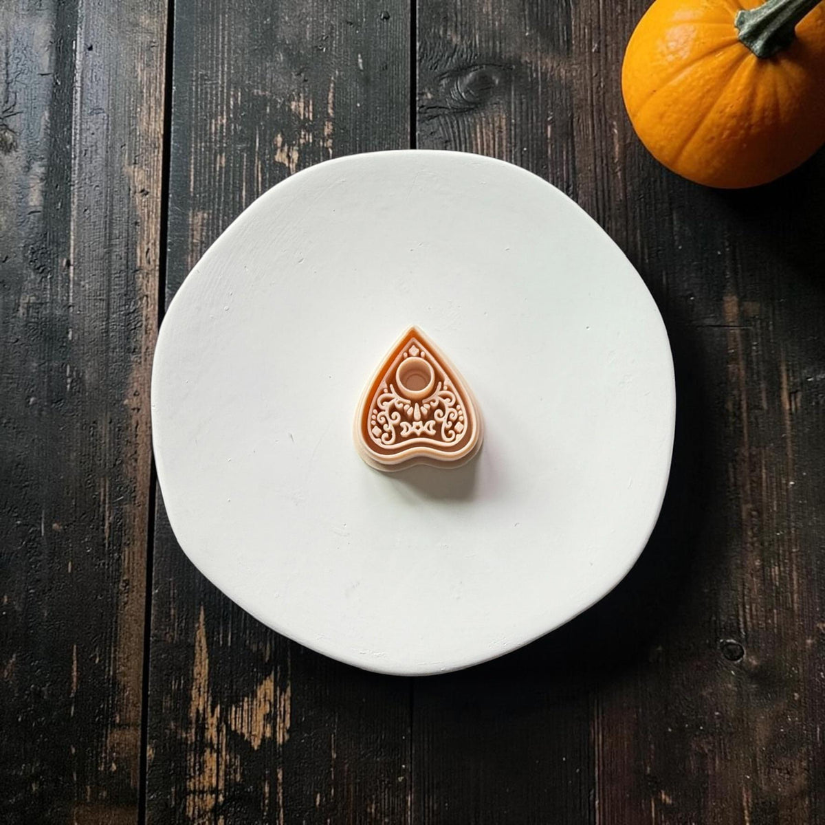 Peach planchette polymer clay cutter centered on a white plate, featuring a pointed shape, circular opening, crescent moon, celestial accents, and ornate decorative scrollwork.