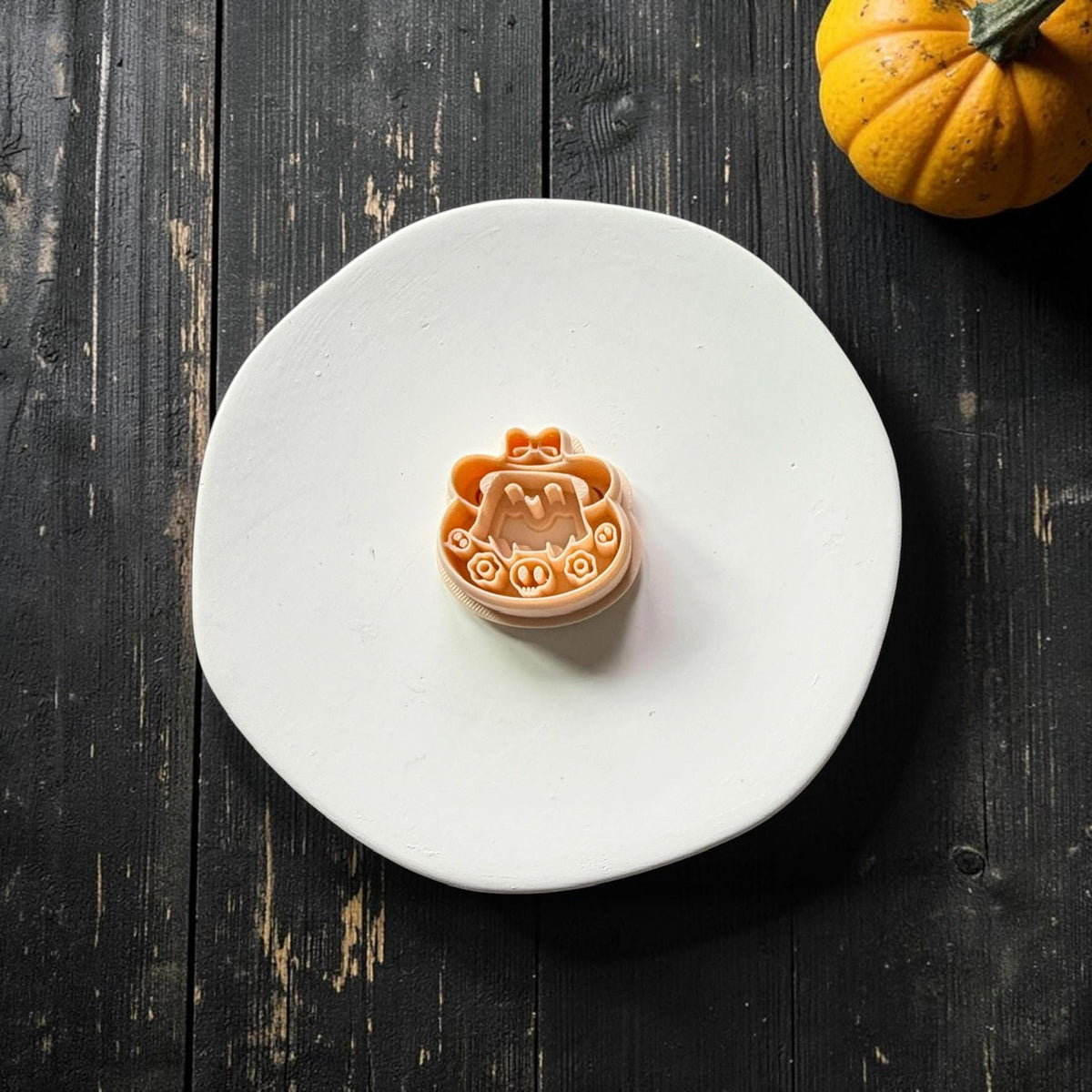 Peach polymer clay cutter featuring a cute smiling ghost beneath an oversized hat with a bow, surrounded by flowers and small skull decorations.