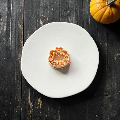 Peach polymer clay cutter featuring a cute smiling ghost beneath an oversized hat with a bow, surrounded by flowers and small skull decorations.