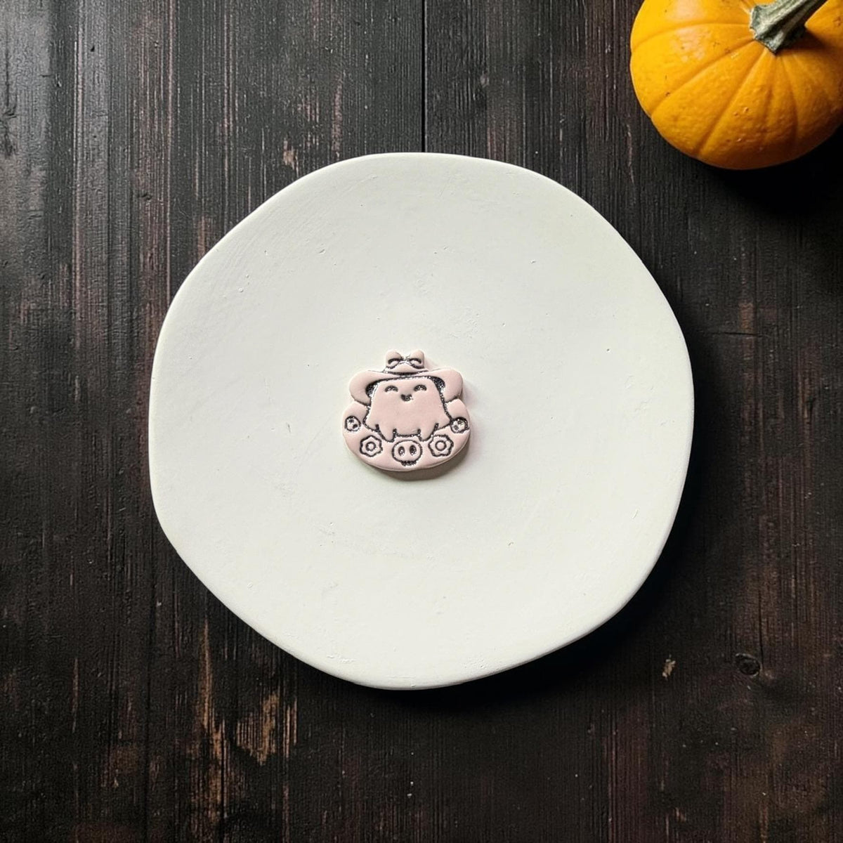 Finished pale pink kawaii ghost clay piece with black accent details, featuring a smiling ghost beneath a large bow-topped hat surrounded by flowers and tiny skulls.