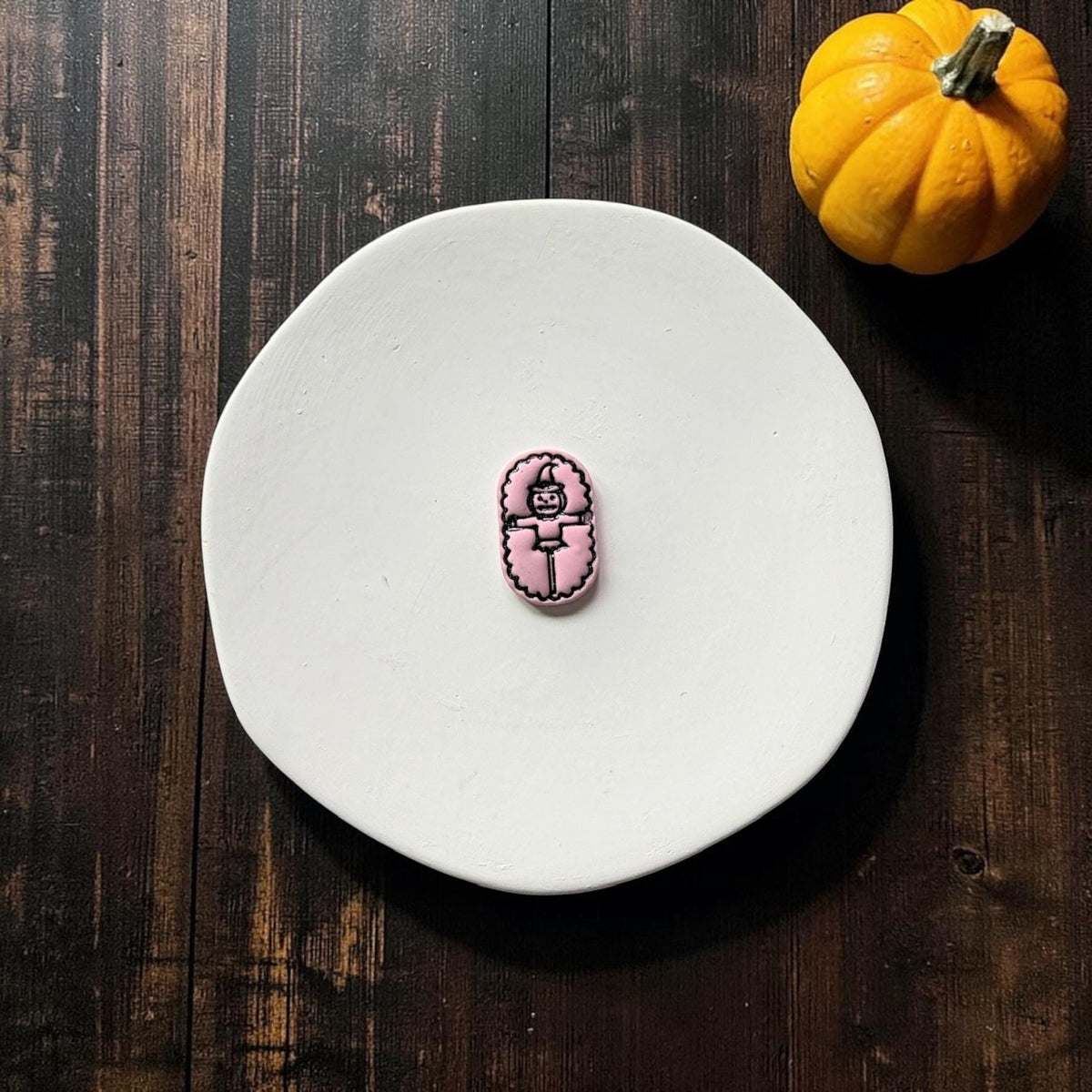 Finished pink pumpkin scarecrow clay piece with black outlined details centered on a white round surface against a dark wood Halloween backdrop.